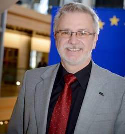 Lächelnder Mann mit Brille, grauem Haar und Bart, im Anzug und roter Krawatte, mit verschwommener EU-Flagge im Hintergrund.