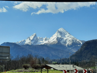 Schneebedeckte Watzmann-Berge unter blauem Himmel mit weißen Wolken, bewaldete Hügel und ein Gebäude im Vordergrund.