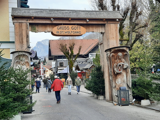 Hölzerner Torbogen mit "Grüß Gott in St. Wolfgang" Schild, geschnitzten Engeln und Menschen auf einer festlichen Dorfstraße.