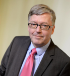 Porträt eines lächelnden Mannes mittleren Alters mit Brille, dunklem Anzug, hellblauem Hemd und pinker Krawatte.