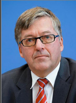 Porträt eines Mannes mittleren Alters mit Brille, hellem Haar, Anzug und rot gestreifter Krawatte vor blauem Hintergrund.