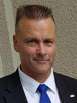 Lächelnder Mann mit kurzen, stacheligen Haaren, dunklem Anzug, gestreiftem Hemd und blauer Krawatte, blickt in die Kamera.