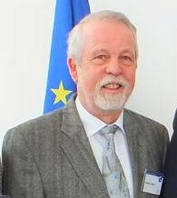 Lächelnder Mann mit grauem Haar und Bart, Anzug, Krawatte, vor einer EU-Flagge.