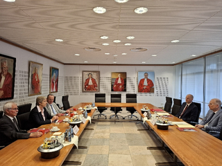Konferenzraum mit sechs Personen an einem U-förmigen Tisch, zahlreiche Porträts von Richtern in roten Roben an den Wänden.