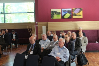 Männer verschiedenen Alters nehmen an einer Konferenz oder einem Seminar teil, sitzend in einem modernen Raum mit großem Fenster und abstrakter Kunst.