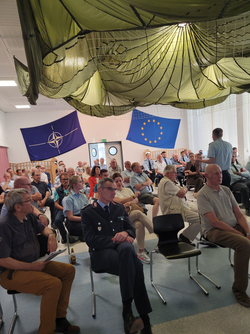 Anwesende, darunter Uniformierte, lauschen einem Redner in einem Raum mit einem Fallschirm an der Decke und NATO- sowie EU-Flaggen.