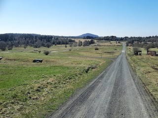 Ein Schotterweg schlängelt sich durch grüne und trockene Felder zu einem entfernten runden Hügel unter blauem Himmel.
