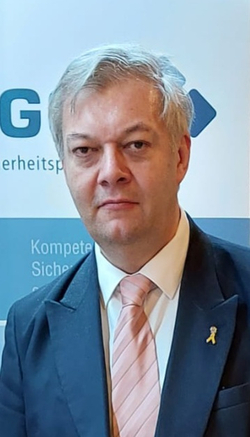 Porträt eines Mannes mit grauem Haar, Anzug, rosa Krawatte und gelber Schleife am Revers.