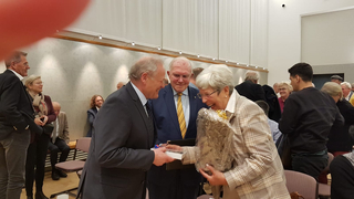 Ein Mann überreicht einer lachenden Frau ein Geschenk, während ein zweiter Mann daneben lächelt, bei einer Veranstaltung.