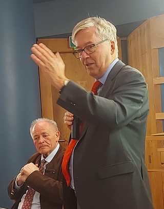 Ein älterer Mann im Anzug und roter Krawatte spricht in ein Mikrofon, gestikulierend, während ein anderer Mann zuhört.