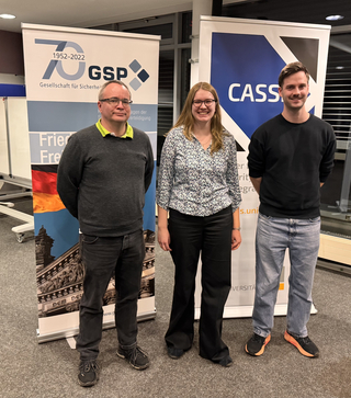 Drei lächelnde Personen, zwei Männer und eine Frau, stehen vor Bannern der Gesellschaft für Sicherheit und CASS Universität.