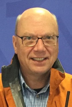 Lächelnder Glatzkopf mit Brille in oranger Jacke hinter einem Rednerpult mit Mikrofonen vor blauer Wand mit Muster.