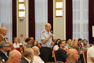 Ein Mann im hellblauen Hemd spricht in ein Mikrofon vor einem sitzenden Publikum in einem Konferenzraum.