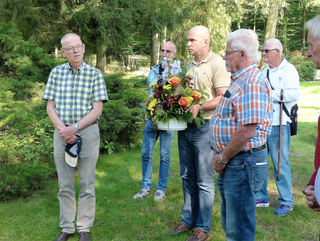 Männergruppe im Grünen, einer hält einen großen Blumenstrauß mit bunten Blumen. Sie stehen auf einer Wiese vor Bäumen.