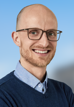 Porträt eines lächelnden, glatzköpfigen Mannes mit Brille und Bart, blauem Hemd und Pullover, Arme verschränkt. Unscharfer Hintergrund.