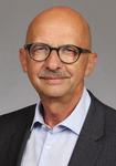 Porträt eines glatzköpfigen Mannes mittleren Alters mit Schnurrbart und Brille, in blauem Hemd und dunklem Sakko.