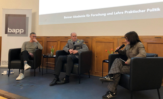 Drei Podiumsgäste, darunter ein Mann in Uniform und eine Frau mit Mikrofon, bei einer Veranstaltung der Bonner Akademie.