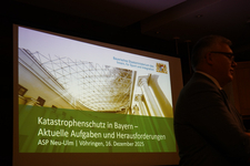 Präsentationsfolie des bayerischen Innenministeriums über Katastrophenschutz in Bayern; ein Redner ist unscharf zu sehen.