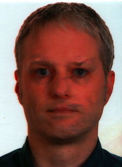 Kopfaufnahme eines Mannes mit kurzen hellbraunen Haaren und blauen Augen, der nach vorne blickt. Das Bild hat einen deutlichen Rotstich.