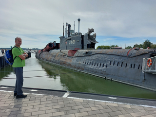 Ein Mann in grünem Shirt und mit Rucksack steht auf einem Kai neben einem großen, rostigen grauen U-Boot U 461 unter bewölktem Himmel.