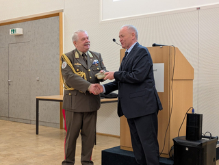 Ein lächelnder Offizier in Uniform und ein Mann im Anzug schütteln Hände und tauschen ein kleines Objekt bei einer Feierlichkeit.