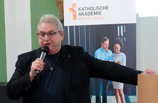 Mann mit Brille spricht in ein Mikrofon bei einer Veranstaltung, mit einem Banner der „KATHOLISCHE AKADEMIE BISTUM FULDA“ im Hintergrund.