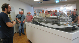 Ein bärtiger Mann spricht zu einer Gruppe, die ein detailliertes Modell eines modernen Kreuzfahrtschiffes in einer Vitrine betrachtet.