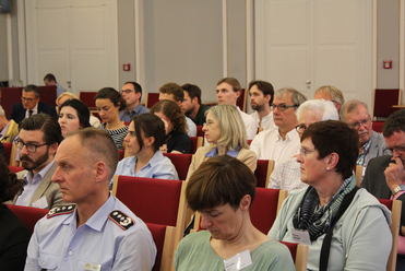 Zuhörer verschiedener Altersgruppen sitzen in einem Konferenzraum mit roten Sitzen und verfolgen eine Veranstaltung.