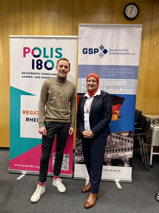 Mann und Frau lächeln vor Bannern von Polis 180 (Grassroots Think Tank) und GSP (Gesellschaft für Sicherheitspolitik).
