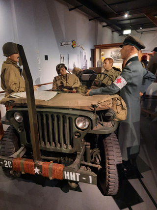 WK2-Museumsausstellung mit vier Militärmannequins und einem Jeep. Ein Sanitäter steht bei, während andere Karten auf der Motorhaube ansehen.
