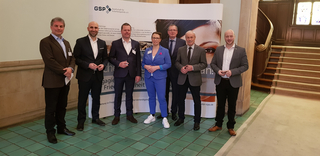 Sieben Personen (sechs Männer, eine Frau) posieren lächelnd vor einem GSP (Gesellschaft für Sicherheitspolitik) Banner.