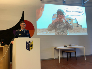 Militäroffizier am Rednerpult mit Wappen, ein Projektor zeigt einen Soldaten in taktischer Ausrüstung, der 'Gibt es noch Fragen?' fragt.