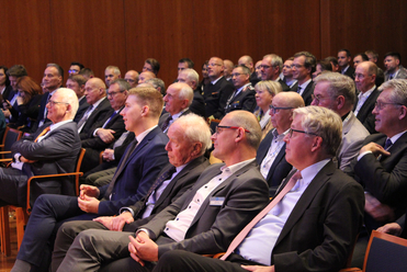 Zahlreiche Teilnehmer in Anzügen sitzen aufmerksam im Publikum einer Business-Veranstaltung oder Konferenz.