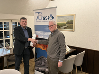Zwei Männer schütteln sich bei einer GSP-Jubiläumsveranstaltung die Hände. Ein Banner für Sicherheitspolitik, Frieden und Freiheit ist im Hintergrund.