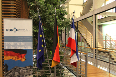 Flaggen der Europäischen Union, Deutschlands und Frankreichs neben einem GSP-Banner 'Frieden sichern Freiheit bewahren' in einem modernen Gebäude.