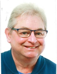Lächelnder Mann mit kurzen grauen Haaren und Brille, der ein blaues Oberteil trägt.