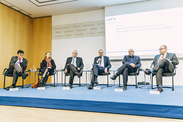 Sechs Podiumsteilnehmer, darunter eine Frau, sitzen auf einer Bühne während einer Konferenz. Ein Mann spricht ins Mikrofon.