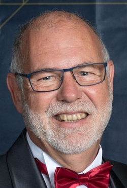 Porträt eines lächelnden, glatzköpfigen Mannes mit grauem Bart, blauer Brille und roter Fliege.