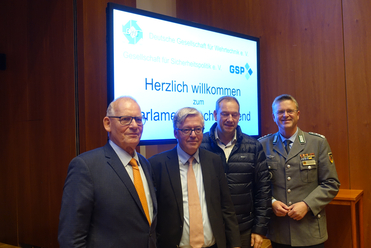 Vier Männer, darunter einer in Militäruniform, stehen vor einem Bildschirm mit 'Herzlich willkommen zum Parlamentsabend'.