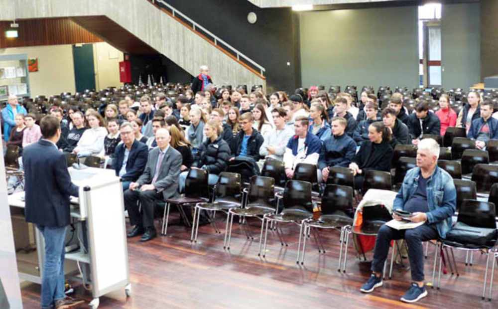 Ein Redner am Pult spricht zu einem großen Publikum, hauptsächlich jungen Leuten, in einem Auditorium bei einer Präsentation.