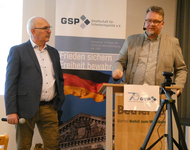 Zwei Männer bei einer GSP-Veranstaltung: Einer spricht am Rednerpult, der andere hält ein Mikrofon. Banner im Hintergrund.