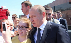 Wladimir Putin steht mit einer jungen Frau, die ein Selfie macht, umgeben von Menschen im Freien.