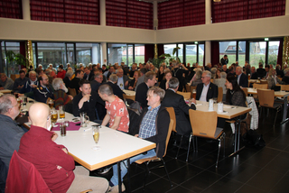 Eine große Gruppe von Erwachsenen und Senioren sitzt in einem Saal an Tischen, unterhält sich, isst und trinkt.