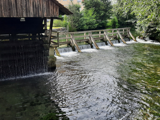 Altes Holzgebäude an einem Fluss mit mehrteiligem Wehr, das kleine Wasserfälle erzeugt, umgeben von grünen Bäumen.