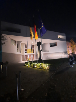 Modernes Gebäude bei Nacht mit wehenden deutschen, EU- und gelben Flaggen. Eine große „25“ ist am beleuchteten Eingang sichtbar.