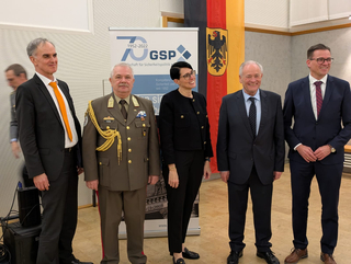 Fünf Personen, darunter ein Offizier, lächeln vor einem GSP 70-Jahre-Banner und der deutschen Flagge.
