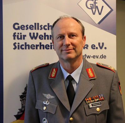 Lächelnder Mann in deutscher Militäruniform mit Medaillen und Namensschild 'WARNECKE' vor einem 'GfW'-Banner.