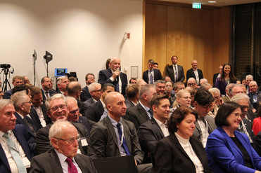 Ein glatzköpfiger Mann mit Bart spricht in ein Mikrofon vor einem sitzenden Publikum bei einer Konferenz.
