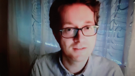 Ein junger Mann mit Brille und kurzen braunen Haaren lächelt leicht und blickt in die Kamera. Er trägt ein helles Hemd vor blauem Hintergrund.