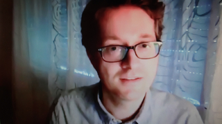 Ein junger Mann mit Brille und kurzen braunen Haaren lächelt leicht und blickt in die Kamera. Er trägt ein helles Hemd vor blauem Hintergrund.